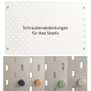 May include: Four different colored plastic knobs for covering screws on a white pegboard. The pegboard has the text "Schraubenabdeckungen für Ikea Skadis" printed on it.