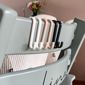 Puede incluir: Un conjunto de cuatro ganchos en colores blanco, beige y negro están unidos a la parte posterior de una silla verde claro. La silla tiene el nombre "Lina" escrito en la parte posterior en letras negras.