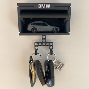 Peut inclure: Un porte-clés noir en forme de garage avec le mot "BMW" en haut. Un modèle de voiture grise est à l'intérieur du garage. Des clés sont suspendues en dessous. Le porte-clés est fixé sur un mur blanc.