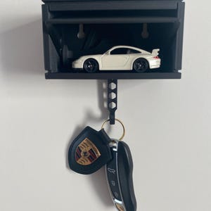 Peut inclure: Un porte-clés mural noir avec une voiture jouet blanche à l'intérieur. Le porte-clés a un crochet pour accrocher les clés. Un porte-clés en cuir noir avec un logo Porsche doré est accroché au crochet.