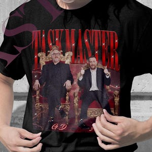 Może przedstawiać: Różowe i czarne koszulki z czerwonym napisem "TASKMASTER" i grafiką dwóch mężczyzn na tronach. Różową koszulkę nosi osoba z blond warkoczami i okularami. Czarną koszulkę nosi osoba z blond włosami.