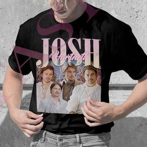 Puede incluir: Dos camisetas, una rosa y otra negra, con un diseño vintage que muestra el texto "Josh Hartnett" y un grupo de hombres. La camiseta rosa la lleva una persona con trenzas rubias, y la camiseta negra la lleva una persona con pelo rubio.