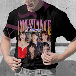 May include: A light pink t-shirt and a black t-shirt, each featuring a graphic of a group of women with the text "Constance Zimmer" in a retro style. The shirts are worn by two people.