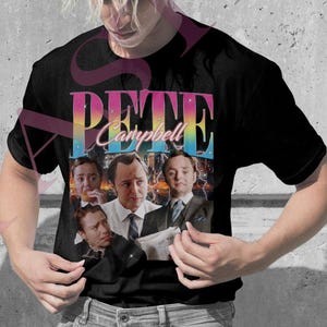 Può includere: Due magliette, una rosa e una nera, ciascuna con una grafica di un uomo e il nome "Pete Campbell" in un font retrò colorato. La maglietta rosa è indossata da una persona con trecce bionde, e la maglietta nera è indossata da una persona con capelli biondi a spillo.