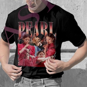 Puede incluir: Camisetas rosas y negras con un grupo de personas con trajes rojos. La palabra "PEARL" está impresa encima de la imagen en una fuente estilizada. Las camisetas las llevan dos personas delante de un fondo gris.