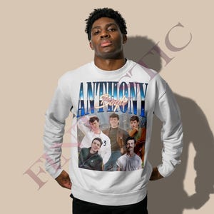 May include: White sweatshirt featuring a group of people with the text "ANTHONY" in a blue and white gradient font. The sweatshirt has a vintage style design with a photo collage of the group. The long-sleeved crew neck sweatshirt is a casual style.