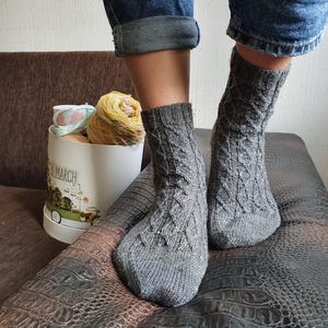 May include: A pair of gray knitted socks with a cable knit pattern. The socks are worn by a person wearing blue jeans.