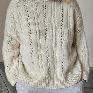 May include: A cream-coloured knitted sweater with cable and openwork detailing. The sweater has a ribbed hem and cuffs, and a crew neck. The design features vertical cable knit panels interspersed with openwork stripes. The sleeves are long and the fit is relaxed.