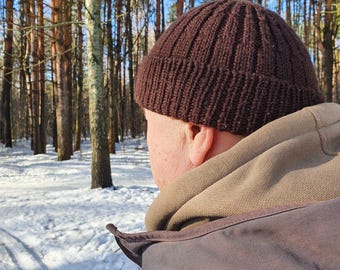 May include: A person wearing a brown knitted beanie in a snowy forest setting.