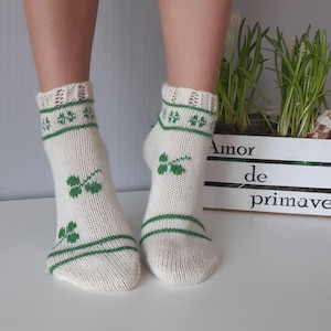 Puede incluir: Un par de calcetines de punto blanco roto con diseños de hojas de trébol verdes. Los calcetines tienen rayas verdes en la parte superior e inferior. El texto "Amor de primavera" es visible en el fondo.