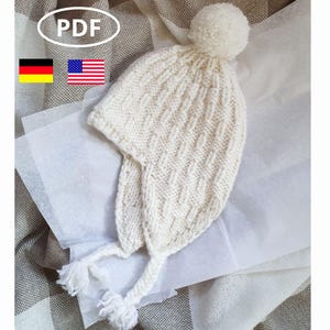 Könnte beinhalten: Weiße Strickmütze mit Bommel oben und langem Quaste. Die Mütze liegt auf einer weißen Oberfläche.