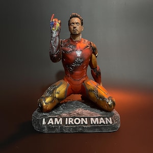 K&ouml;nnte beinhalten: Eine rote und goldene Actionfigur von Iron Man, die kniet und den rechten Arm erhoben hat, wobei sie den Infinity Gauntlet mit allen sechs Infinity Stones h&auml;lt. Die Figur steht auf einem schwarzen Steinsockel mit der Aufschrift "I AM IRON MAN".