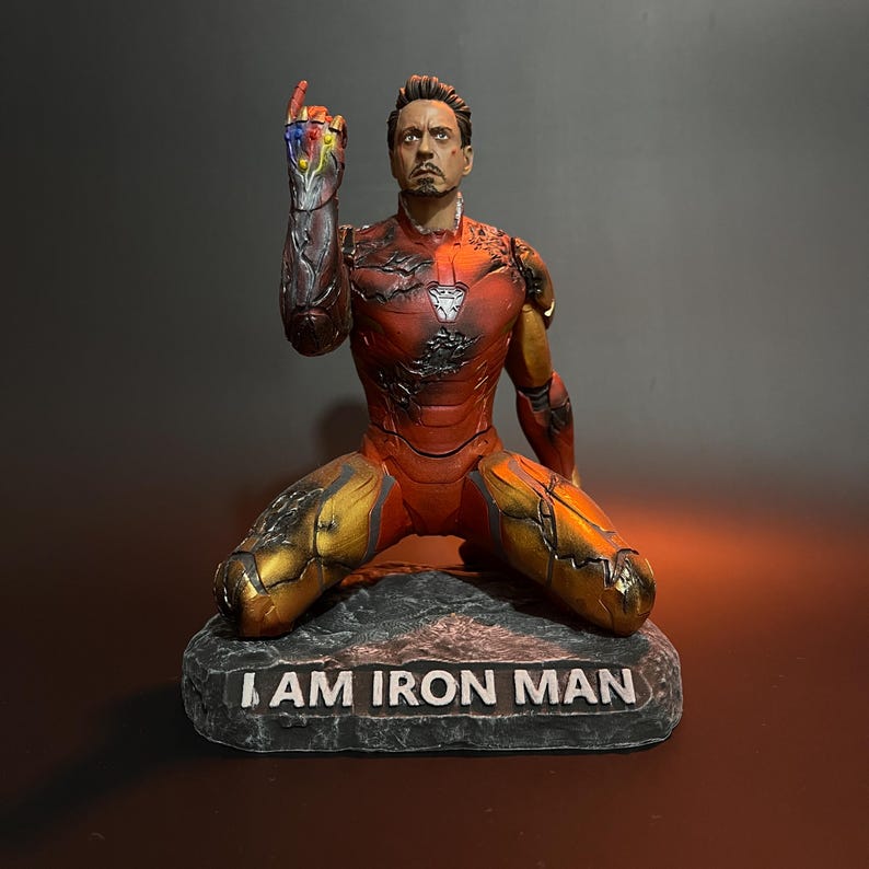K&ouml;nnte beinhalten: Eine rote und goldene Actionfigur von Iron Man, die kniet und den rechten Arm erhoben hat, wobei sie den Infinity Gauntlet mit allen sechs Infinity Stones h&auml;lt. Die Figur steht auf einem schwarzen Steinsockel mit der Aufschrift "I AM IRON MAN".