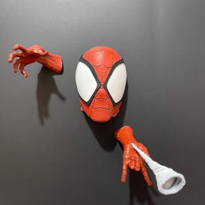 Puede incluir: Una máscara de Spider-Man roja y negra con ojos blancos y un diseño de telaraña. Dos manos de Spider-Man rojas y negras están unidas a la pared, una con una telaraña blanca.