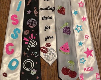 May include: Five satin neckties featuring various designs: one white with pink and blue glitter text 'KISS', one black with disco balls and 'POP' text, one white with 'it's all waiting there for you', one grey with glitter fruit, and one white with stars and text.