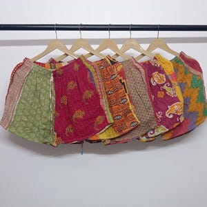 Op de afbeelding: Een verzameling kleurrijke, bedrukte shorts die aan houten hangers hangen. De shorts hebben verschillende ontwerpen, waaronder bloemen- en geometrische patronen, in tinten rood, groen, geel en bruin. De shorts zijn gemaakt van een lichte stof.