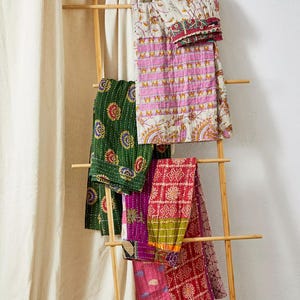 May include: A wooden ladder displays a collection of colourful, patterned textiles. The textiles include a green piece with floral designs, a pink and white piece with geometric patterns, and a red piece with a patchwork design. The textiles are draped over the ladder.