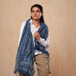 Peut inclure: Écharpe à motifs bleu marine et blancs avec des franges. Elle est portée sur une chemise blanche et un pantalon beige. Le motif présente un motif de chevrons répétés. L'ensemble est décontracté et élégant.