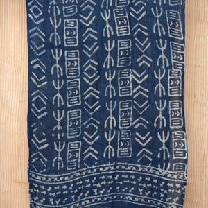 Peut inclure: Une écharpe bleu marine avec un motif géométrique blanc et des franges. L'écharpe est faite d'un tissu léger et présente un motif répétitif de rectangles, de losanges et d'autres formes. L'écharpe est suspendue sur un fond brun clair.