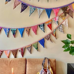 Könnte beinhalten: Eine bunte, handgefertigte Tragetasche mit Patchwork-Design liegt auf einem braunen Sofa. Darüber hängen zwei Girlanden aus dreieckigen Stoffwimpeln in verschiedenen Mustern und Farben an einer beigen Wand. Ein rotes Tablett mit Kerzen und einem dekorativen Objekt befindet sich im Vordergrund.