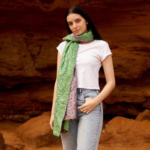 May include: A green and purple scarf with a textured pattern. The scarf is draped around the neck and shoulders. The scarf is made of a soft material. The background is a reddish-brown rock formation.