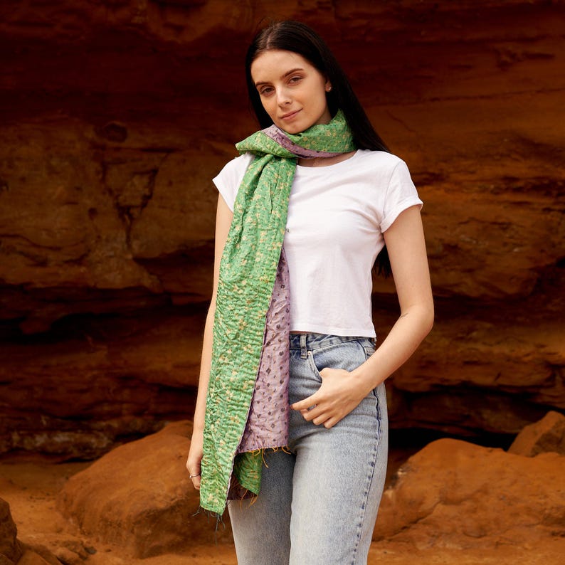 May include: A green and purple scarf with a textured pattern. The scarf is draped around the neck and shoulders. The scarf is made of a soft material. The background is a reddish-brown rock formation.