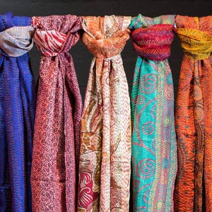 Könnte beinhalten: Eine Sammlung farbenfroher Schals, jeder mit einzigartigen Mustern und Texturen. Die Schals sind über eine Stange drapiert und zeigen eine Vielzahl von Farben, darunter Blau, Rosa, Orange, Türkis und Gelb. Die Stoffe scheinen leicht und vielseitig einsetzbar.