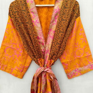 K&ouml;nnte beinhalten: Ein lebendiger, gemusterter Bademantel in Orange-, Braun- und Rosat&ouml;nen, der an einem wei&szlig;en Kleiderb&uuml;gel h&auml;ngt. Der Bademantel hat ein Blumen- und Paisley-Design mit passendem Bindeg&uuml;rtel. Die &Auml;rmel sind weit und der Stoff wirkt leicht.