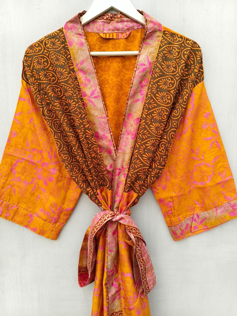 K&ouml;nnte beinhalten: Ein lebendiger, gemusterter Bademantel in Orange-, Braun- und Rosat&ouml;nen, der an einem wei&szlig;en Kleiderb&uuml;gel h&auml;ngt. Der Bademantel hat ein Blumen- und Paisley-Design mit passendem Bindeg&uuml;rtel. Die &Auml;rmel sind weit und der Stoff wirkt leicht.