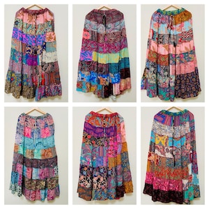 Puede incluir: Seis faldas patchwork coloridas, compuestas por diversos patrones y colores de tela, incluyendo rosa, morado, azul y naranja. Las faldas están colgadas en perchas de madera sobre un fondo blanco. Las faldas tienen un diseño escalonado.