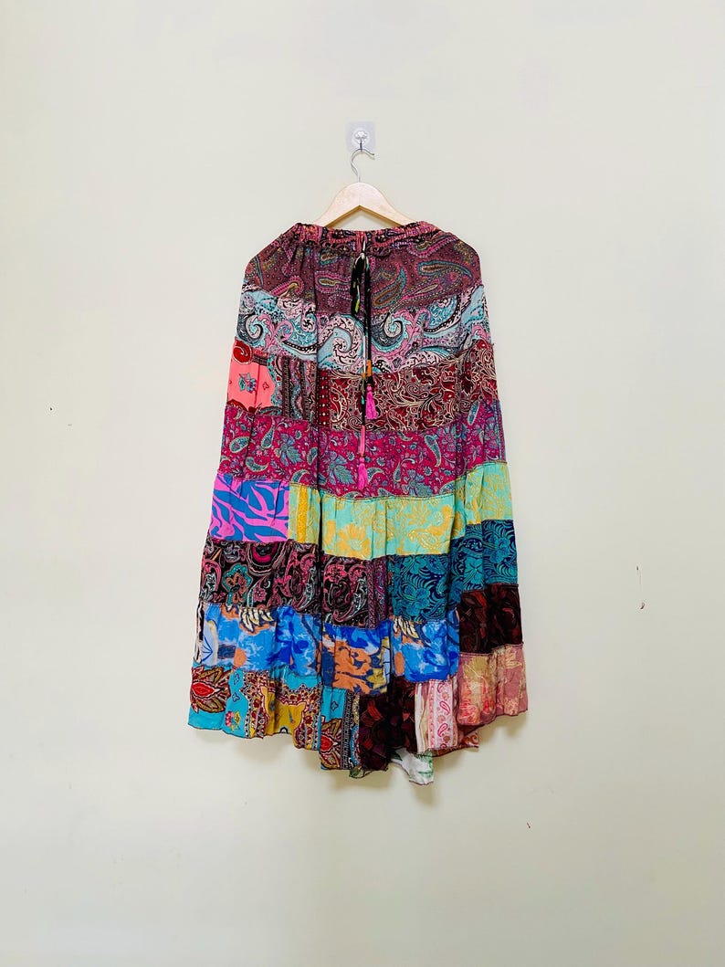 Puede incluir: Una falda patchwork con una variedad de coloridos estampados de cachemira y florales. La falda est&aacute; hecha de m&uacute;ltiples paneles de tela en tonos de rosa, azul, verde y burdeos. Cuelga de una percha de madera.