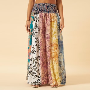 May include: A long, flowing skirt with a patchwork design. The skirt features panels of various colours and patterns, including paisley, floral, and abstract prints. The waistband is a dark blue, gathered fabric. The skirt is worn with sandals.