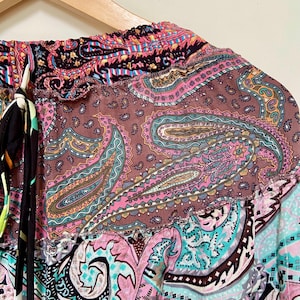 Puede incluir: Primer plano de una prenda con estampado de cachemira colorida. La tela presenta una mezcla de tonos rosa, marr&oacute;n, turquesa y negro. La prenda est&aacute; colgada de una percha de madera, mostrando el intrincado dise&ntilde;o y los colores vibrantes.