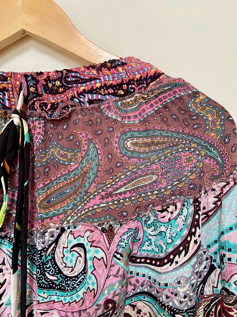 Puede incluir: Primer plano de una prenda con estampado de cachemira colorida. La tela presenta una mezcla de tonos rosa, marr&oacute;n, turquesa y negro. La prenda est&aacute; colgada de una percha de madera, mostrando el intrincado dise&ntilde;o y los colores vibrantes.