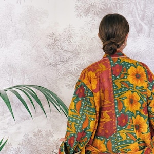 May include: A colorful, floral-patterned robe in shades of yellow, red, and green. The robe features a patchwork design with large flower motifs and a contrasting blue trim. The garment is worn by a person with their back to the camera.