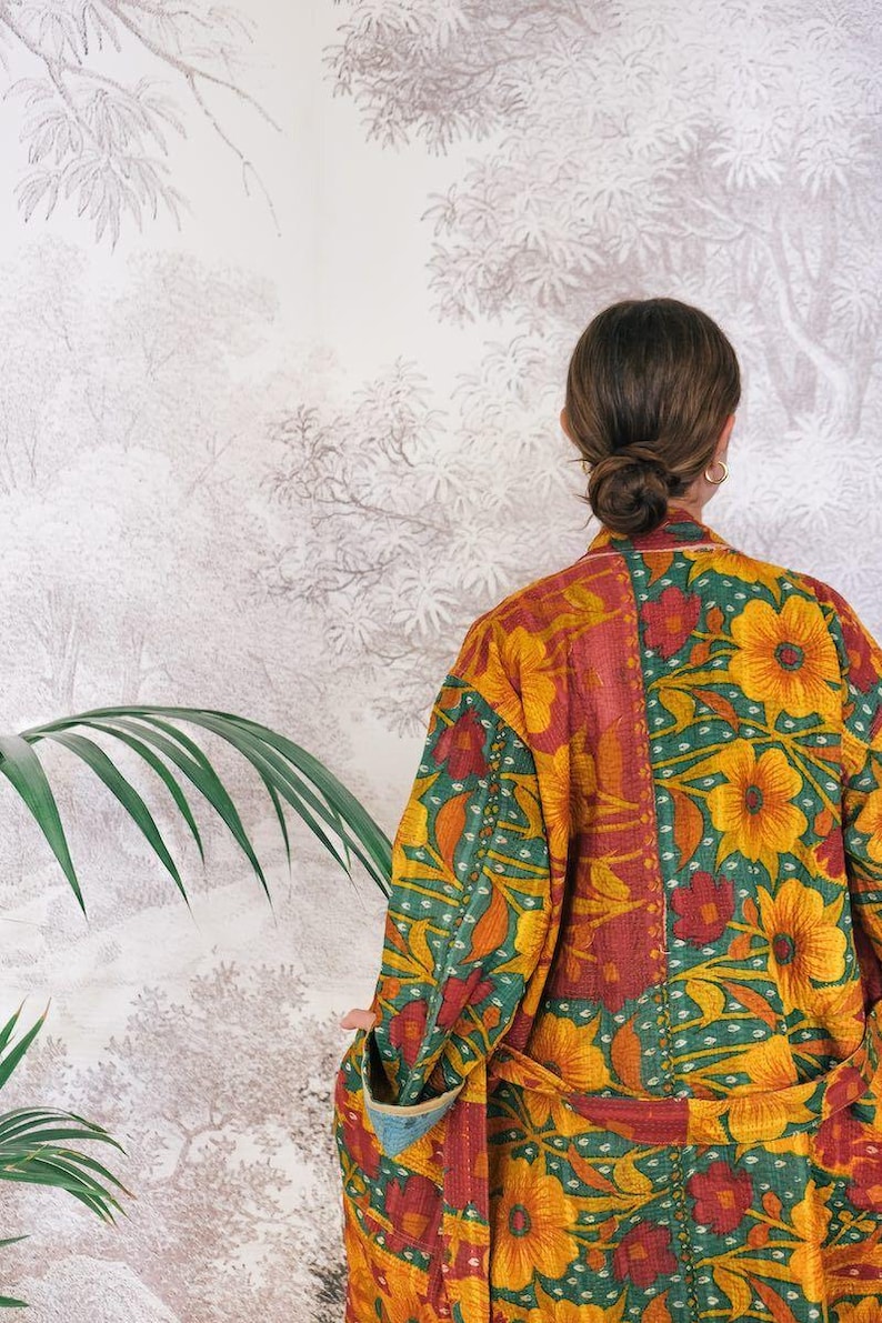 May include: A colorful, floral-patterned robe in shades of yellow, red, and green. The robe features a patchwork design with large flower motifs and a contrasting blue trim. The garment is worn by a person with their back to the camera.