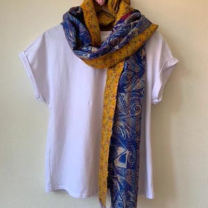 May include: A white t-shirt on a hanger with a colorful scarf draped over it. The scarf features a blue and yellow paisley design with fringed edges. A pink gingham heart decorates the top of the hanger.