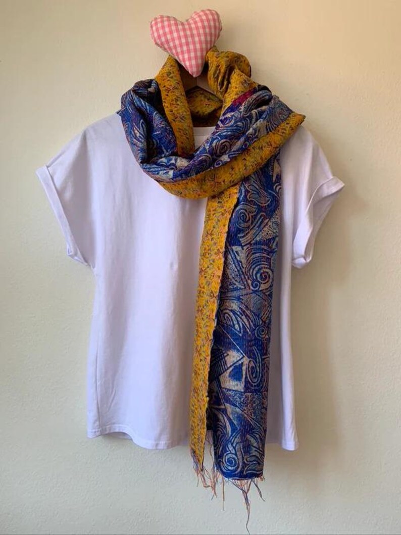 May include: A white t-shirt on a hanger with a colorful scarf draped over it. The scarf features a blue and yellow paisley design with fringed edges. A pink gingham heart decorates the top of the hanger.