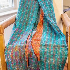 May include: A colorful textile scarf draped over a chair. The scarf features a teal and orange design with intricate patterns and stitching. The fabric has a textured appearance, with visible seams and a fringed edge. The scarf is likely handmade.