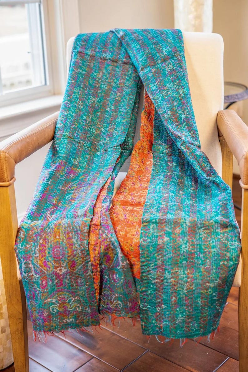 May include: A colorful textile scarf draped over a chair. The scarf features a teal and orange design with intricate patterns and stitching. The fabric has a textured appearance, with visible seams and a fringed edge. The scarf is likely handmade.
