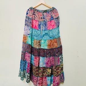 Puede incluir: Una falda larga de patchwork con una variedad de coloridos estampados de cachemira y geom&eacute;tricos. La falda est&aacute; hecha de tela ligera y tiene una cintura con cord&oacute;n. La falda cuelga de una percha de madera sobre un fondo blanco.