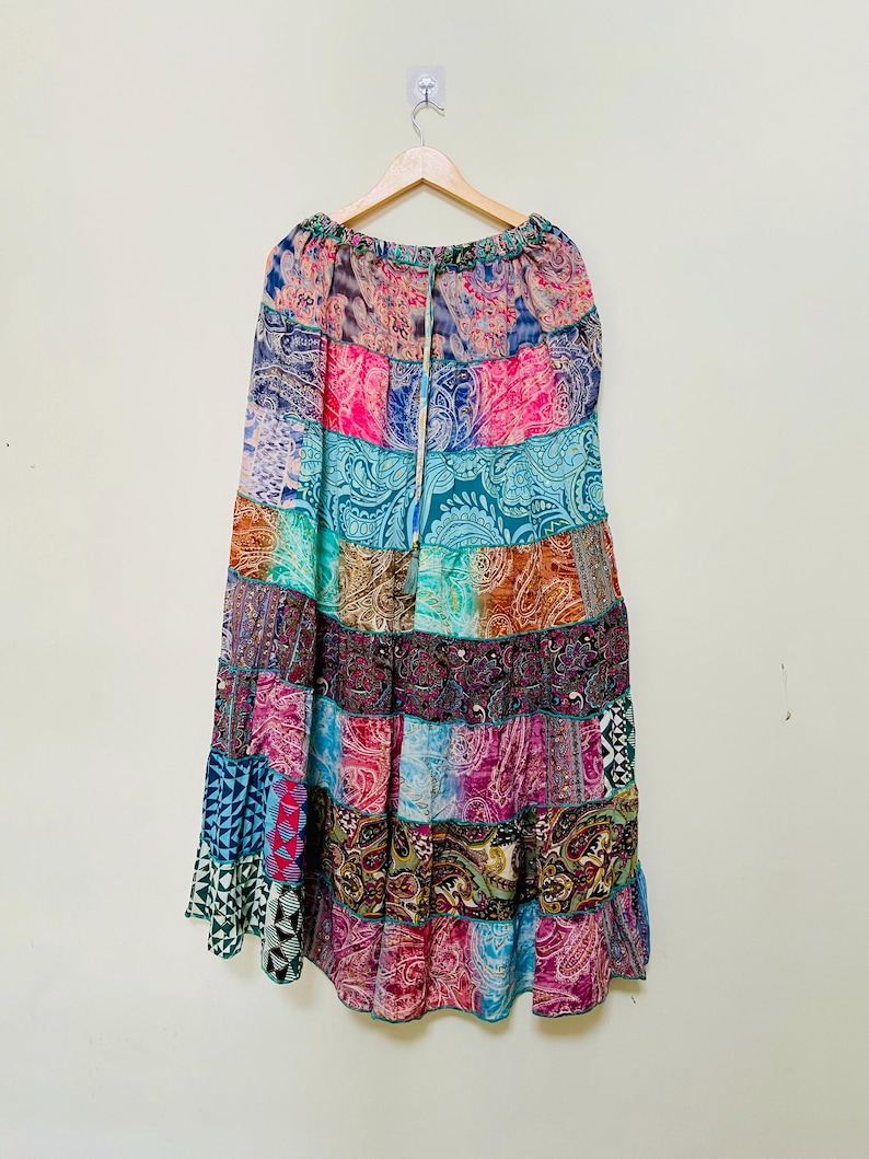 Puede incluir: Una falda larga de patchwork con una variedad de coloridos estampados de cachemira y geom&eacute;tricos. La falda est&aacute; hecha de tela ligera y tiene una cintura con cord&oacute;n. La falda cuelga de una percha de madera sobre un fondo blanco.