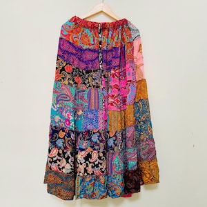 Puede incluir: Una falda larga de patchwork con una variedad de estampados de cachemira en colores vibrantes, incluyendo rosa, azul, morado y naranja. La falda est&aacute; colgada en una percha de madera contra una pared blanca. Tiene cintura con cord&oacute;n.
