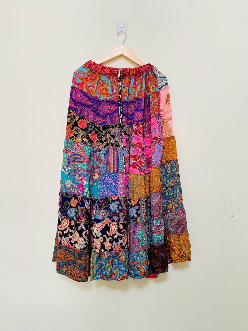 Puede incluir: Una falda larga de patchwork con una variedad de estampados de cachemira en colores vibrantes, incluyendo rosa, azul, morado y naranja. La falda est&aacute; colgada en una percha de madera contra una pared blanca. Tiene cintura con cord&oacute;n.