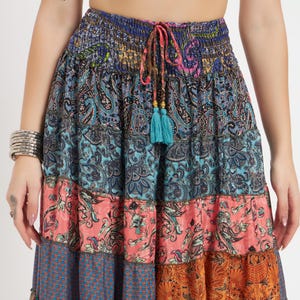 May include: A tiered, bohemian-style skirt with a vibrant mix of patterns and colors. The skirt features paisley, floral, and geometric designs in shades of blue, coral, and orange. A drawstring waist with turquoise tassels adds a decorative touch.