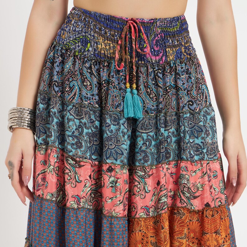 May include: A tiered, bohemian-style skirt with a vibrant mix of patterns and colors. The skirt features paisley, floral, and geometric designs in shades of blue, coral, and orange. A drawstring waist with turquoise tassels adds a decorative touch.