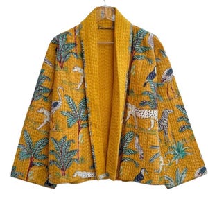 Könnte beinhalten: Senfgelbe Steppjacke mit Dschungel-Print. Die Jacke hat einen Schalkragen und zeigt Illustrationen von Leoparden, Vögeln und Palmen. Sie hängt an einem silbernen Haken vor weißem Hintergrund.