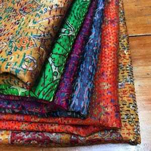 May include: A stack of colorful, patterned fabric pieces. The fabrics feature intricate stitching and a variety of colors, including gold, green, purple, blue, and orange. The designs appear to be floral and geometric.