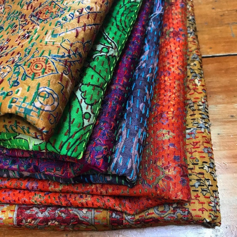 May include: A stack of colorful, patterned fabric pieces. The fabrics feature intricate stitching and a variety of colors, including gold, green, purple, blue, and orange. The designs appear to be floral and geometric.