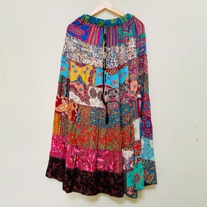 Puede incluir: Una falda larga de patchwork con una variedad de patrones coloridos, incluyendo dise&ntilde;os de cachemira y florales. La falda est&aacute; colgada en una percha de madera sobre un fondo blanco. La falda tiene un cord&oacute;n en la cintura y una borla negra.